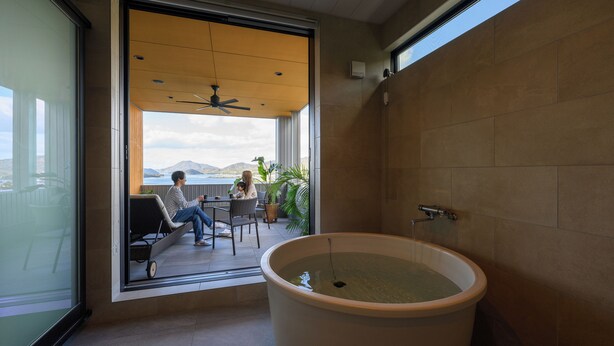 A modern bathroom with a large bathtub, a ceiling fan, and a view of a balcony with two people sitting outside. - Chichibugahama Resort Villa Senja (Mitoyo)