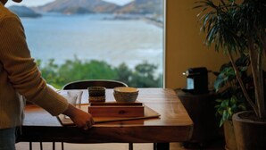 A wooden table with bowls and a glass, a potted plant, and a view of a body of water and mountains. - Chichibugahama Resort Villa Senja (Mitoyo)