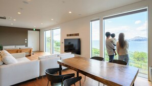 A modern living room with a large TV, a wooden dining table, and a sofa. There are large windows offering a view of the sea. - Chichibugahama Resort Villa Senja (Mitoyo)