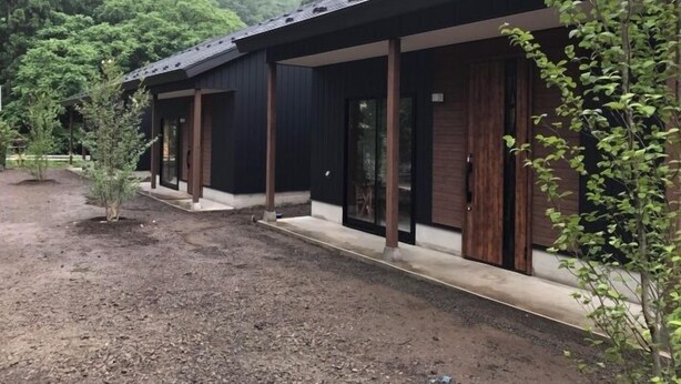 A row of modern, dark-colored buildings with large windows and wooden doors, set in a natural, forested area with a dirt path leading to them.