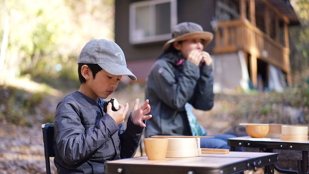 Two individuals seated at an outdoor table with bowls and wooden containers, likely for a meal or activity. - Hutte Lied (Komoro)