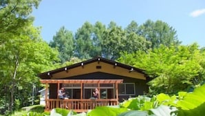 A cabin with a wooden deck and large windows, surrounded by lush greenery. - Hutte Lied (Komoro)