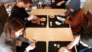 A group of people seated around a table with various items, including cups and plates, suggesting a social gathering or meal. - Hutte Lied (Komoro)