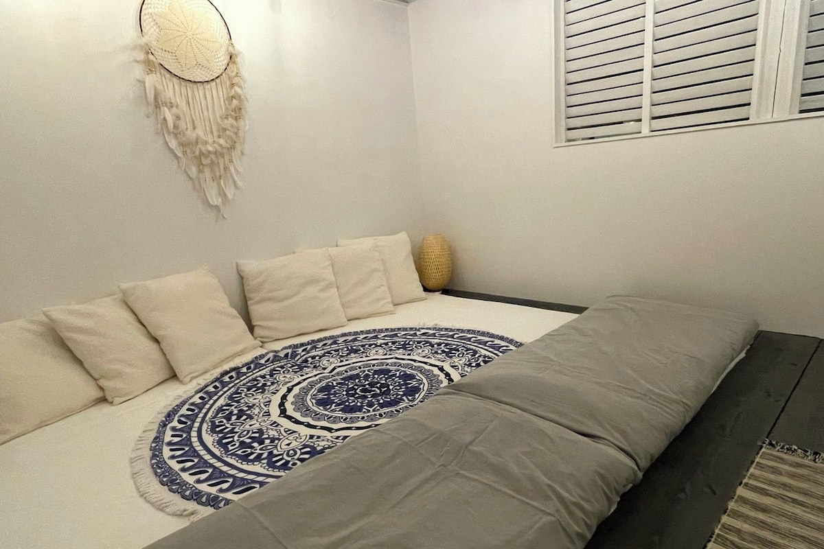 Een slaapkamer met een bed, kussens, een hoofdbord, een aan de muur bevestigde droomvanger, en een raam met jaloezieën.