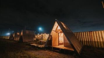 A series of triangular wooden cabins at night, each with a visible interior and a small table outside.