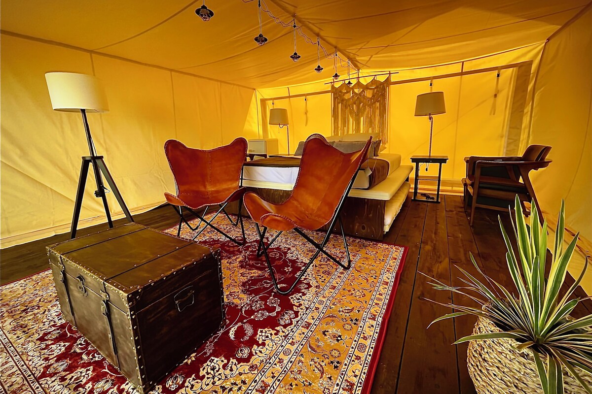 A tented room with a bed, two leather chairs, a small table, a lamp, and a suitcase.