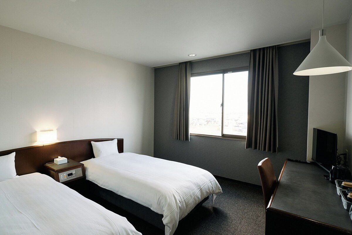A hotel room with two beds, a desk, a chair, a lamp, and a large window with curtains.
