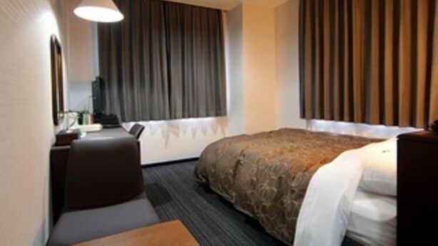 Una habitación de hotel con cama, escritorio, silla, lámpara y ventana con cortinas.