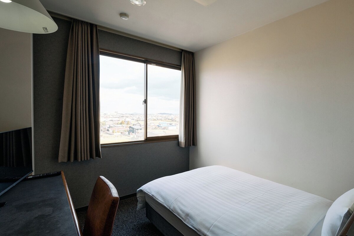 Une chambre d’hôtel avec un lit simple, un bureau, une chaise et une grande fenêtre donnant sur la ville.