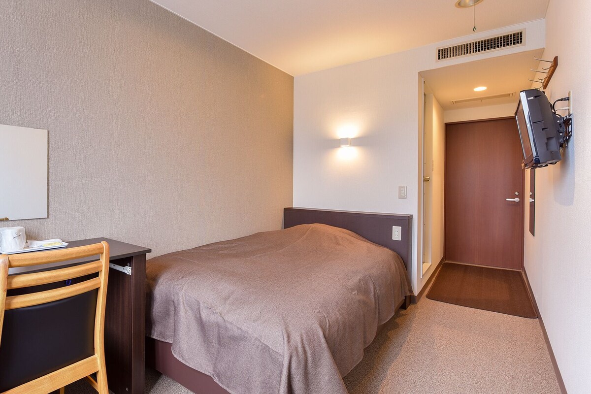 A hotel room with a bed, a desk, a chair, a TV mounted on the wall, and a door leading to another room.