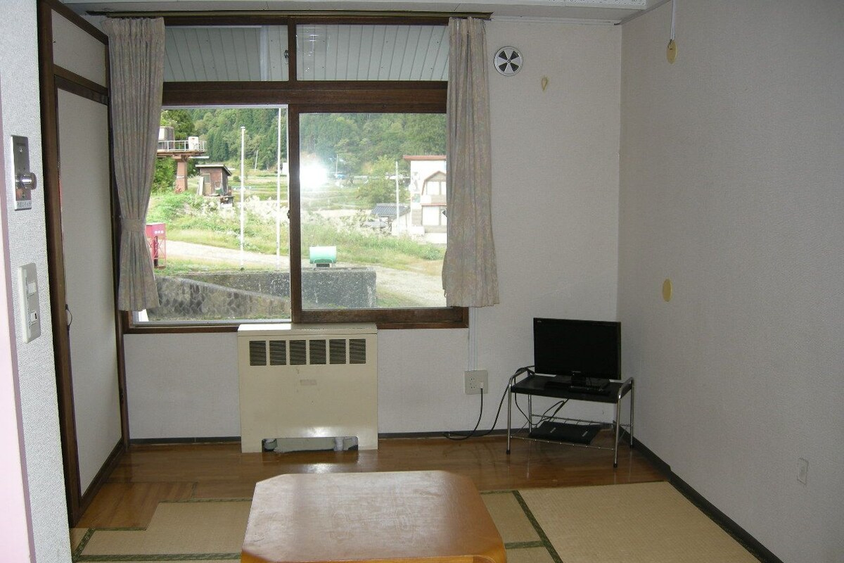 Una habitación con suelo de tatami, una mesa de madera, un televisor sobre un mueble y una ventana con cortinas.