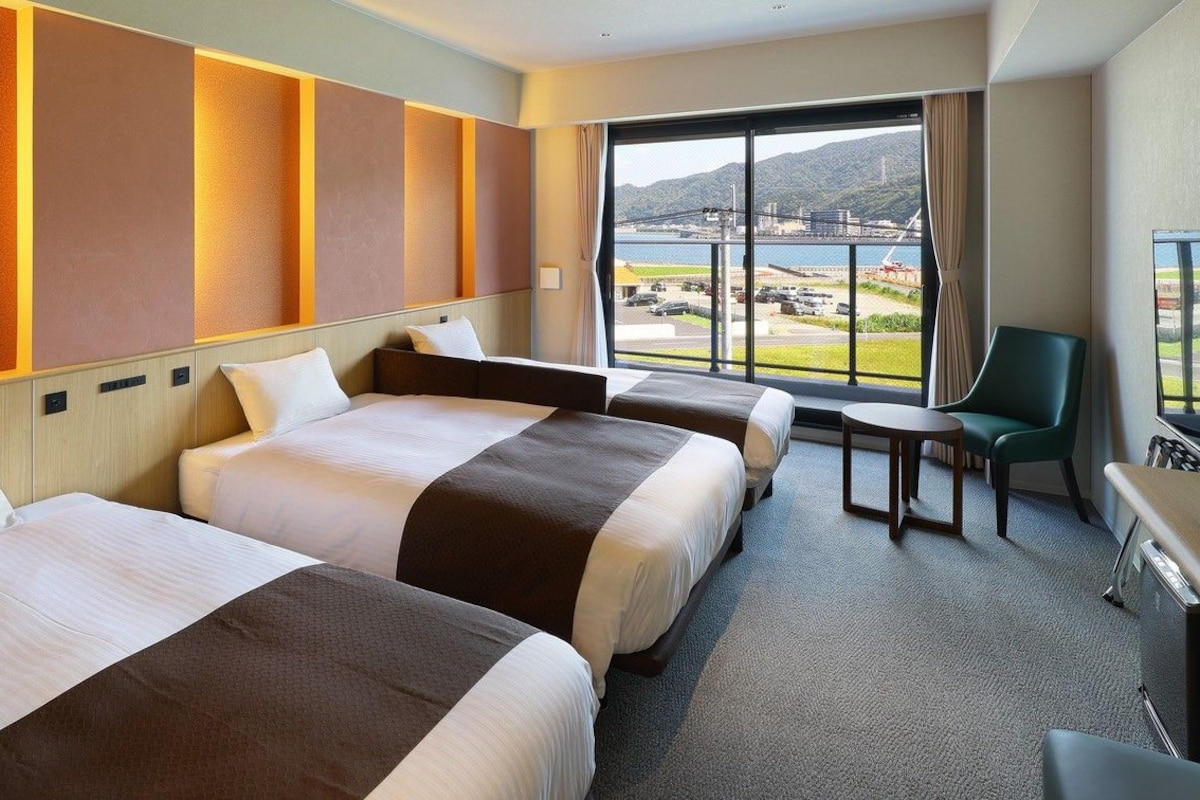 Habitación de hotel con dos camas, una mesita, una silla y un ventanal con vistas al agua y colinas.
