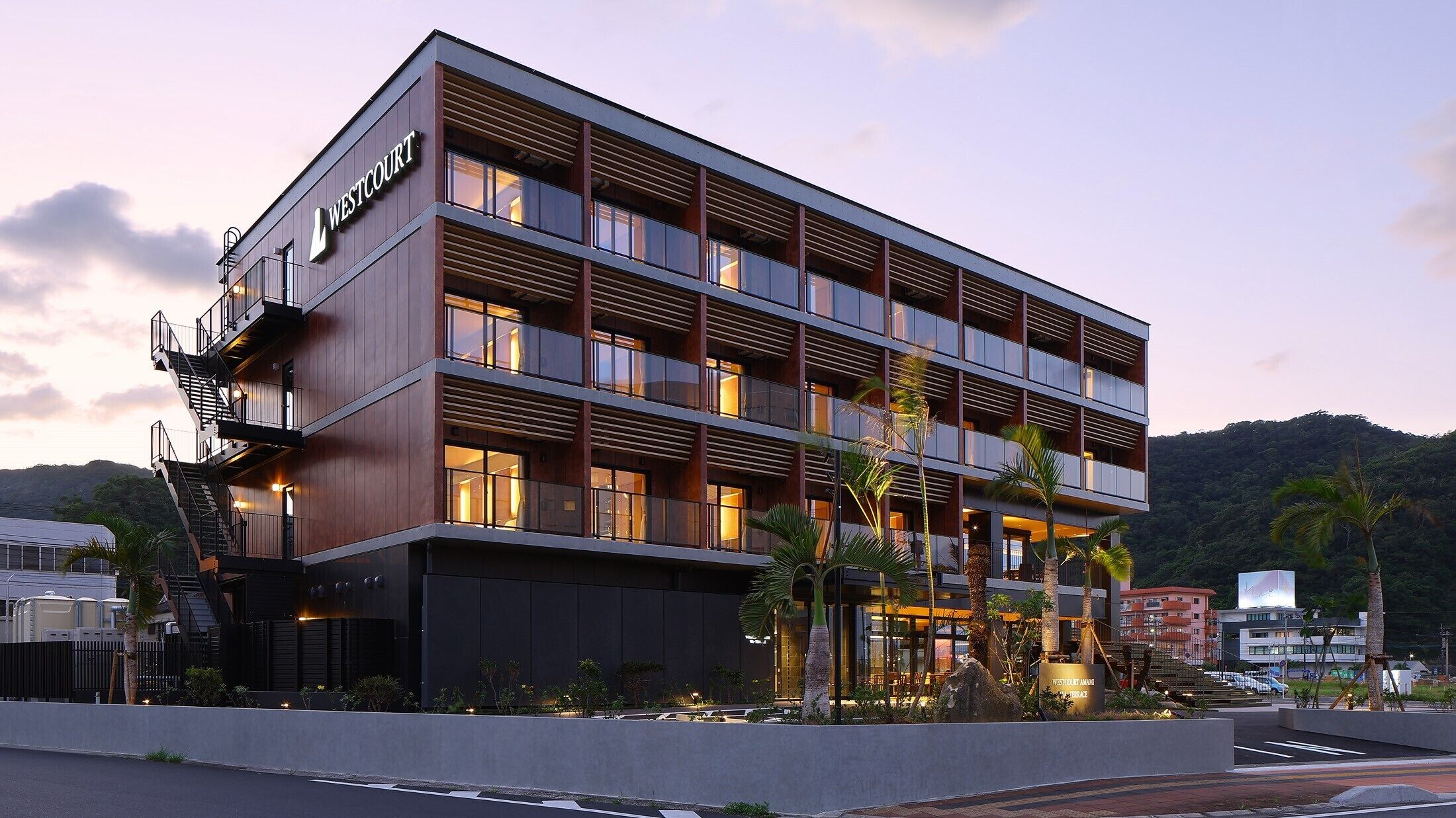 Un hotel de varios pisos con un diseño moderno, que cuenta con balcones en cada planta, una entrada destacada y un área exterior ajardinada con palmeras.