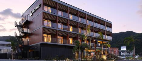 Un hotel de varios pisos con un diseño moderno, que cuenta con balcones en cada planta, una entrada destacada y un área exterior ajardinada con palmeras.