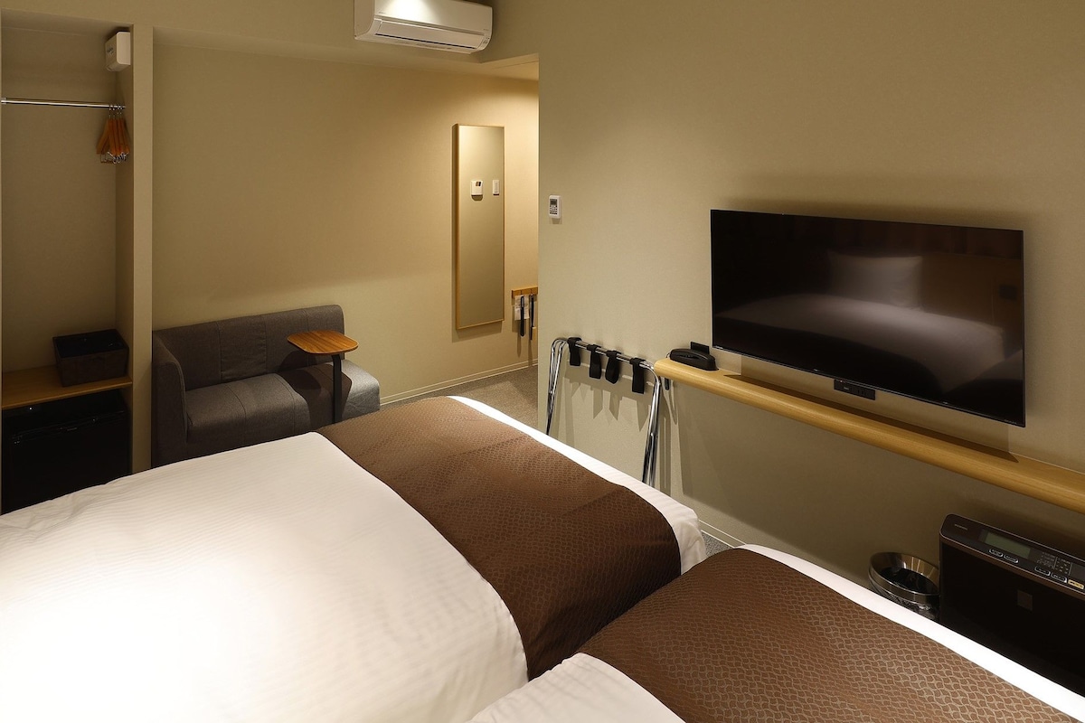Habitación de hotel con dos camas, televisión, escritorio y zona de ducha.