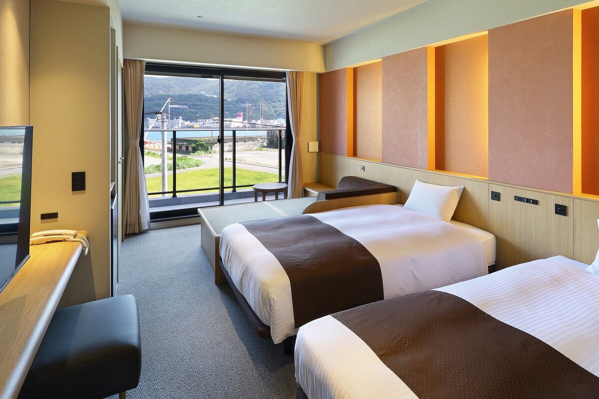Habitación de hotel con dos camas, un escritorio, una silla y un amplio ventanal con vistas a la ciudad y montañas.