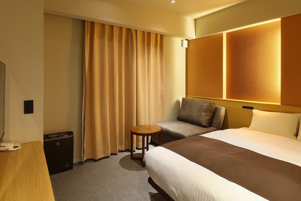 Una habitación de hotel con cama, una mesita, un sofá, un televisor y una cortina.