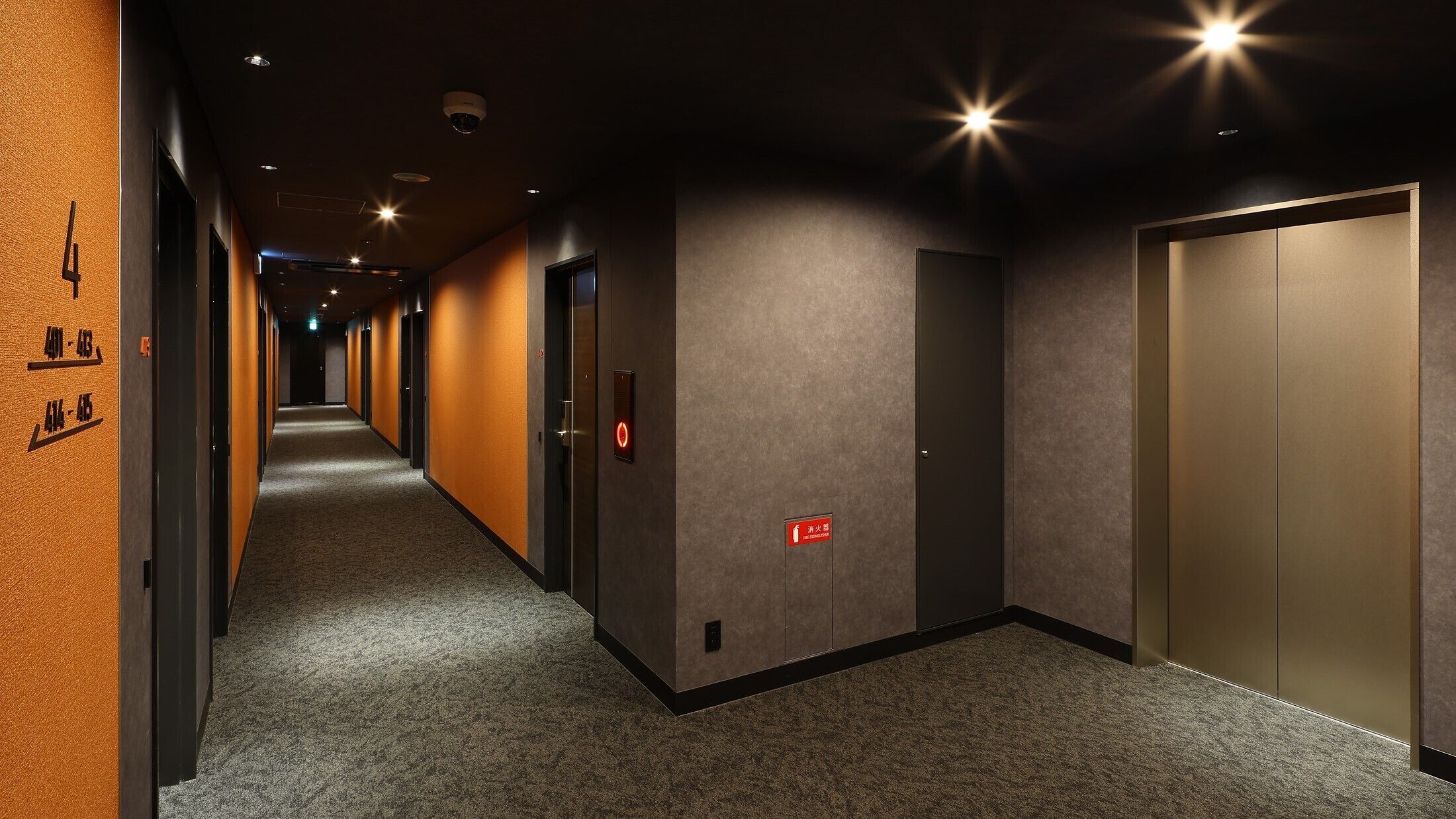 A hotel corridor with a carpeted floor, doors on the left, and a fire exit sign on the right.