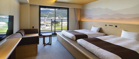 A hotel room with two beds, a sofa, a TV, and a view of a bridge and mountains.