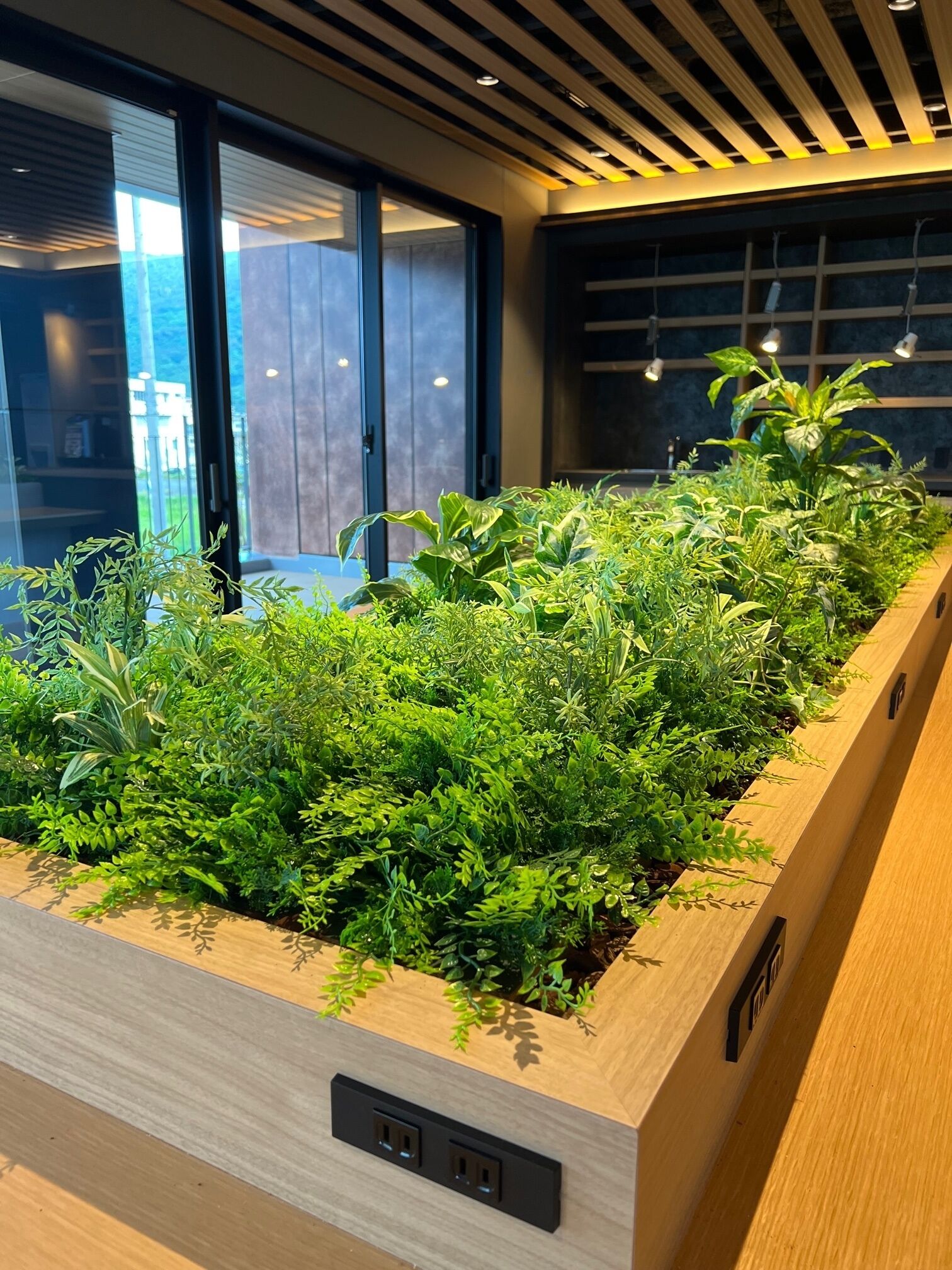 Een moderne binnentuin met een houten plantenbak, ingebouwde stopcontacten en uitzicht op het buitenlandschap door grote ramen.
