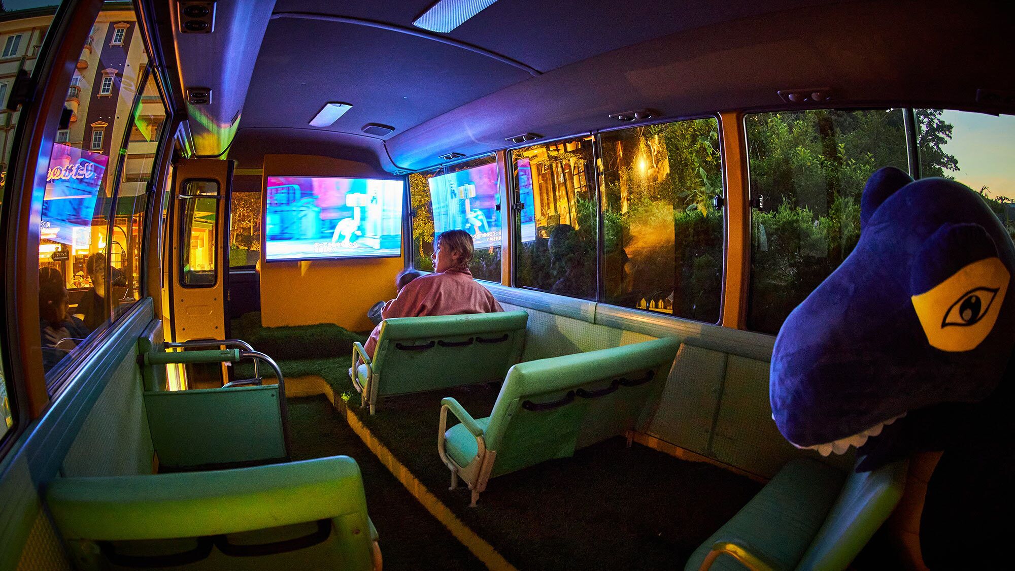 Een businterieur met groene stoelen, een groot scherm waarop een programma wordt vertoond, en een zittende persoon.