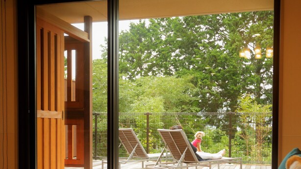 Une pièce avec un plancher en bois, un canapé, une table et une vue sur un balcon où se trouvent une personne et un chien.