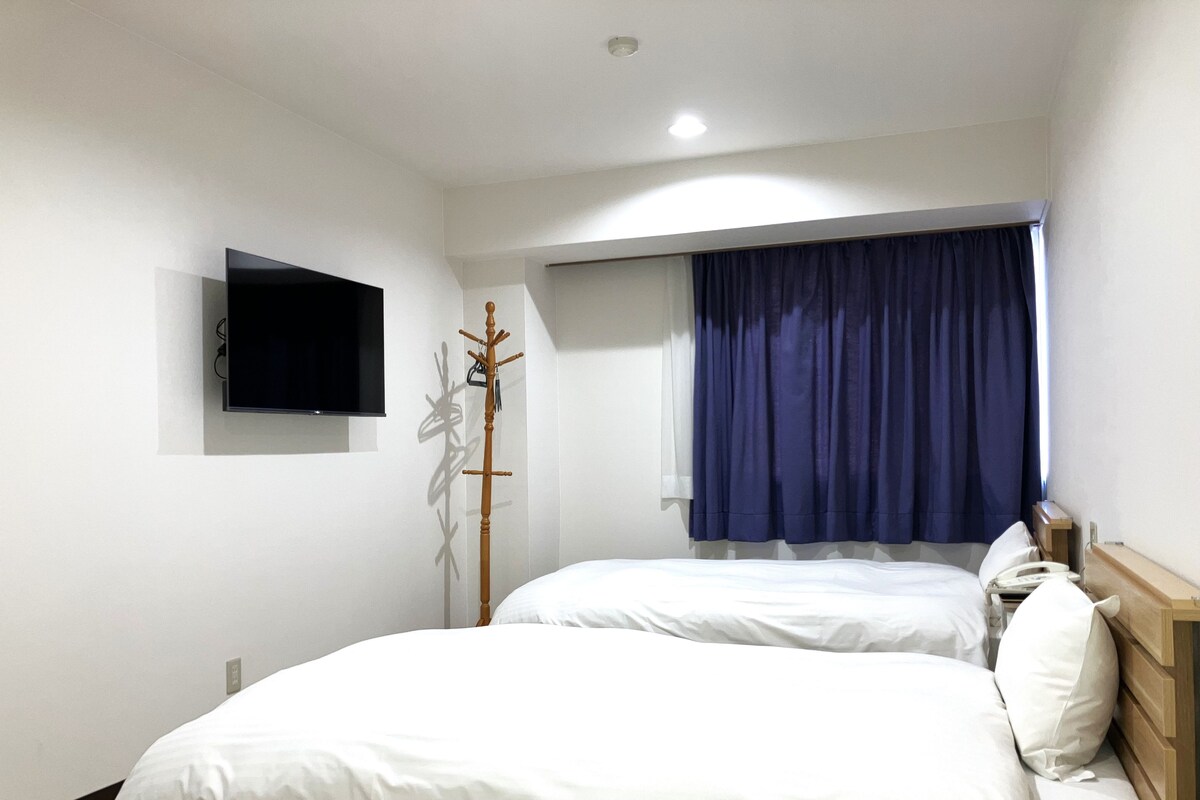 A hotel room with two beds, a TV mounted on the wall, a coat rack, and a window with curtains.