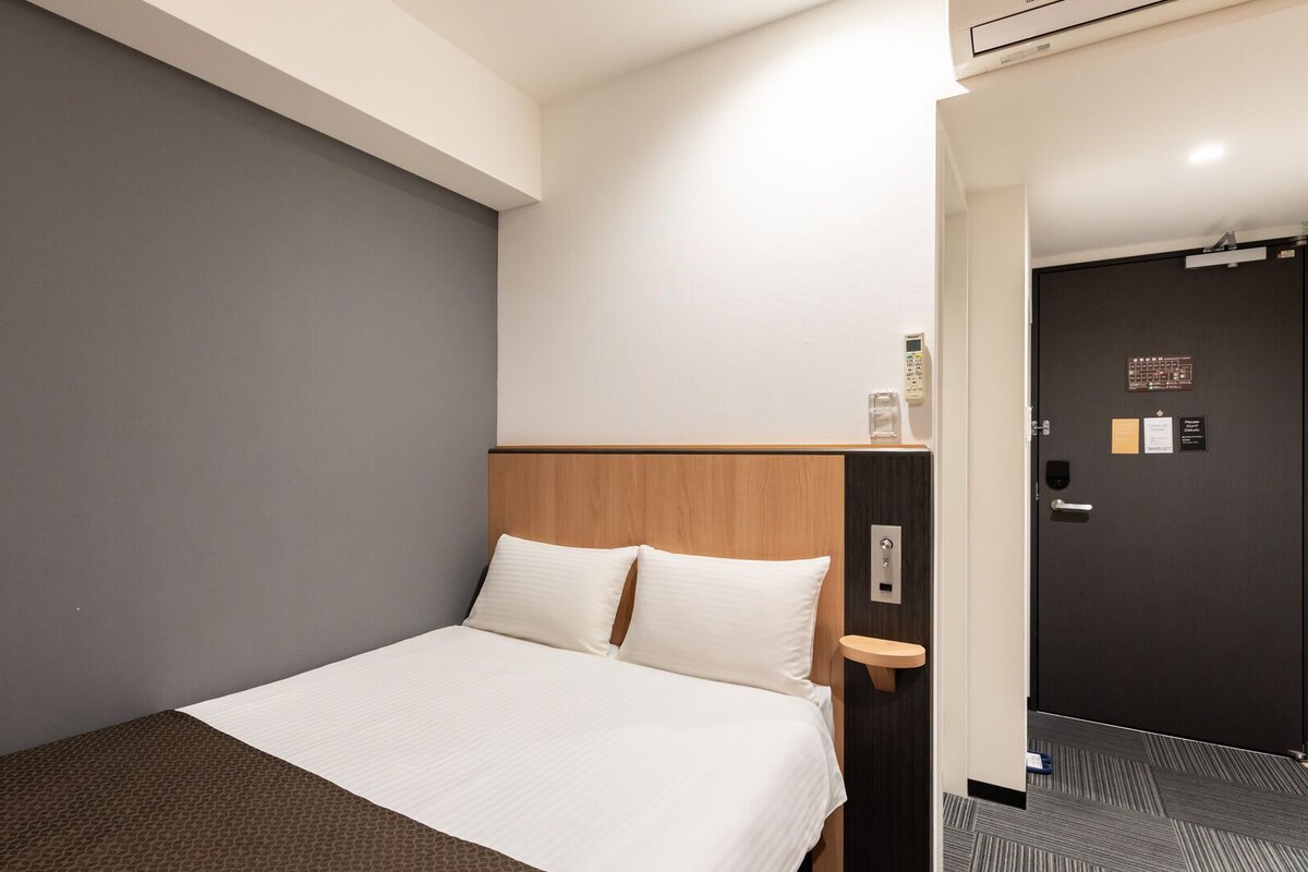 A hotel room with a bed, a bedside table, a wall-mounted phone, and a door with a sign.