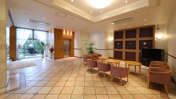 Un hall d’hôtel avec un sol carrelé, un comptoir de réception, un espace détente avec des chaises et des tables, un téléviseur et une grande fenêtre donnant sur un paysage verdoyant.