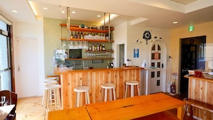 Een bar met houten krukken, een lange houten tafel en planken met flessen en glazen.