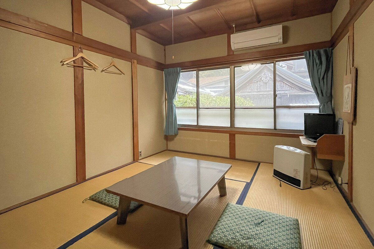 A traditional Japanese room with tatami flooring, a low table, a bench, a desk with a computer, and a view of a traditional building outside.