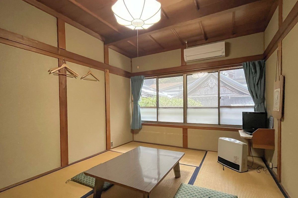 Una habitación japonesa tradicional con suelo de tatami, una mesa baja, un banco, un escritorio con una computadora y vistas a un edificio tradicional exterior.
