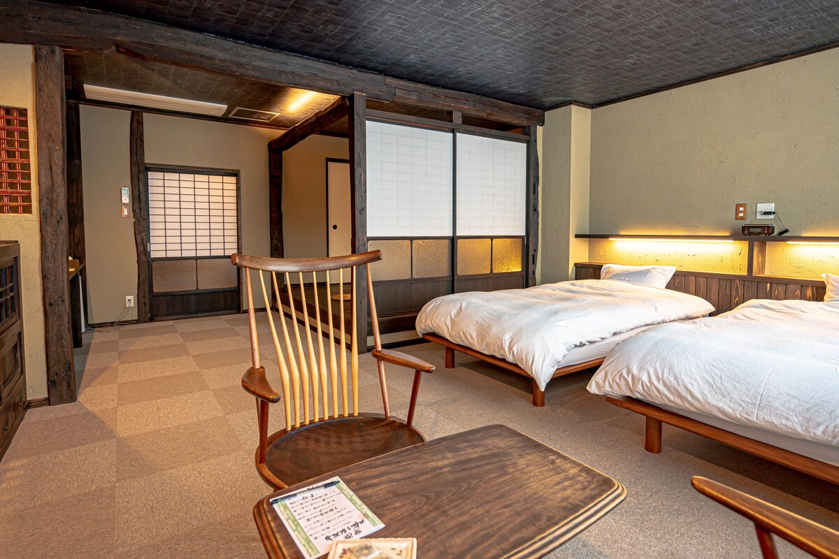 A traditional Japanese-style room with tatami flooring, a low wooden bed, a wooden chair, and a small table with a menu.