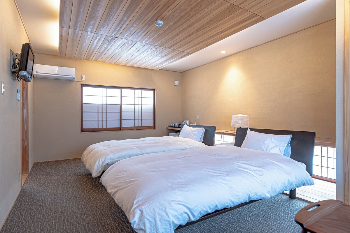 A hotel room with two beds, a large window, a ceiling fan, and a bedside table with a lamp.