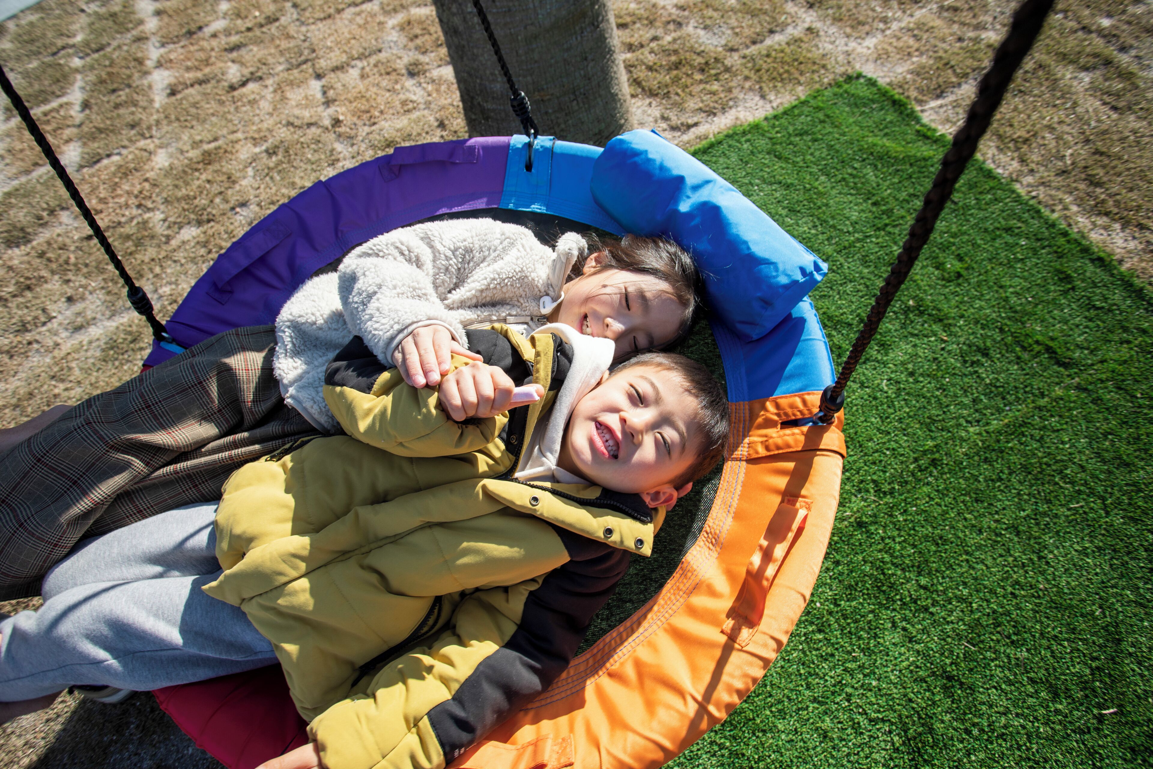 Un enfant et un adulte sont assis sur une balançoire dotée d’un coussin bleu et orange, dans un espace extérieur recouvert de gazon synthétique.