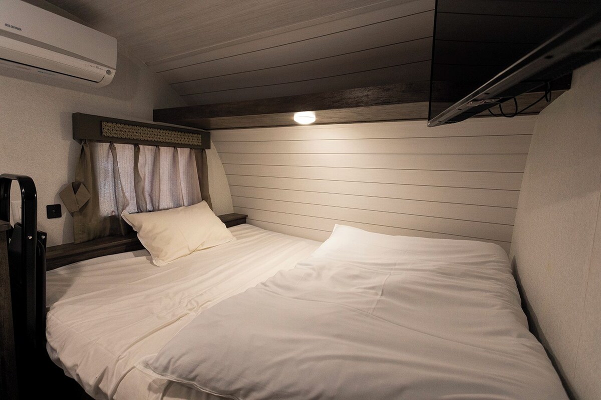Un lit simple avec du linge de lit blanc, dans une pièce aux murs lambrés et dotée d’un luminaire au plafond.