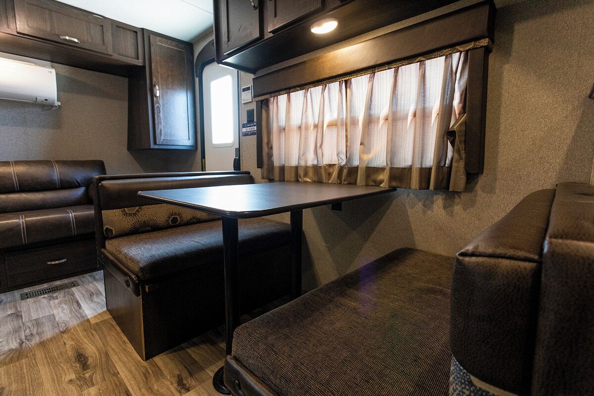 Un intérieur de camping-car compact comprenant une table à manger, des sièges rembourrés et une fenêtre avec des rideaux.