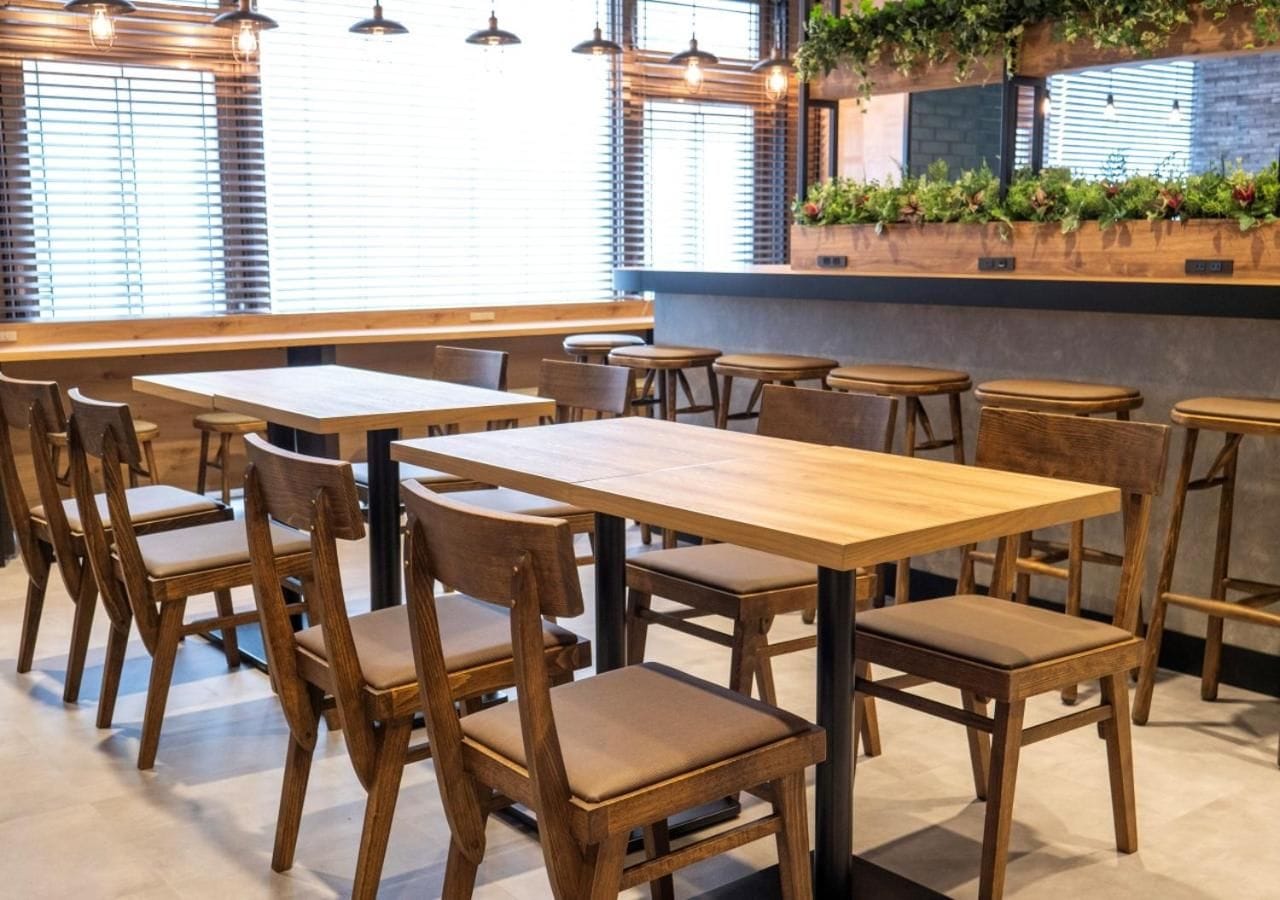 Interior de restaurante con mesas y sillas de madera, una barra con menú y una pared cubierta de plantas.