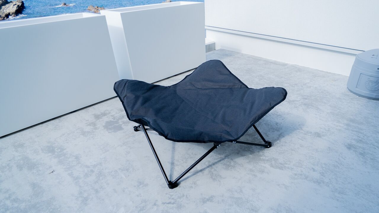 A modern, black, foldable chair with a unique design, placed on a concrete floor near a white wall and a glass window overlooking a body of water.