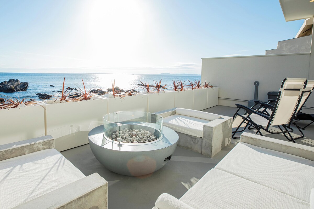 A sunlit outdoor seating area with a glass-top table, white cushioned chairs, and a view of the ocean.