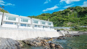 A modern white building with multiple balconies overlooking a rocky shoreline and clear blue water. - Beach Terrace Boso (Futtsu)