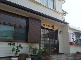Un edificio con un cartel que dice “お食事・民宿” y otro cartel con “小倉会” y un número de teléfono. Hay plantas en macetas al frente.