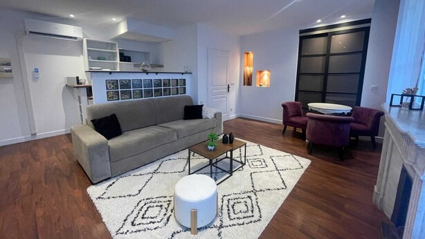 Un salon moderne avec un canapé gris, une table basse et un pouf rond. Une cheminée se trouve à droite, et une grande fenêtre est équipée de stores.