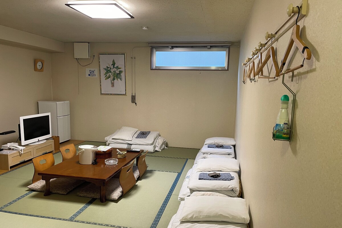 Habitación de hotel con suelo de tatami, una mesa de madera, televisor, refrigerador y ventana con vistas.