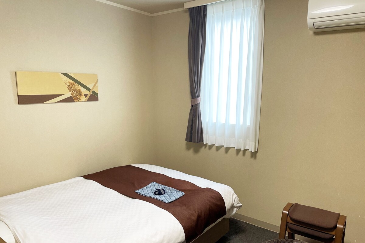 A hotel room with a bed, a chair, a table, a window with curtains, and a wall-mounted air conditioner.