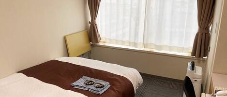A hotel room with a bed, a desk, a chair, a window with curtains, and a small table with a phone and a notepad.