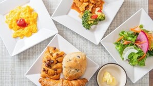 A table with four square plates containing different dishes: scrambled eggs with a cherry, a chicken dish with broccoli, a croissant, and a salad with radish. - Super Hotel Saitama Kuki (Kuki)