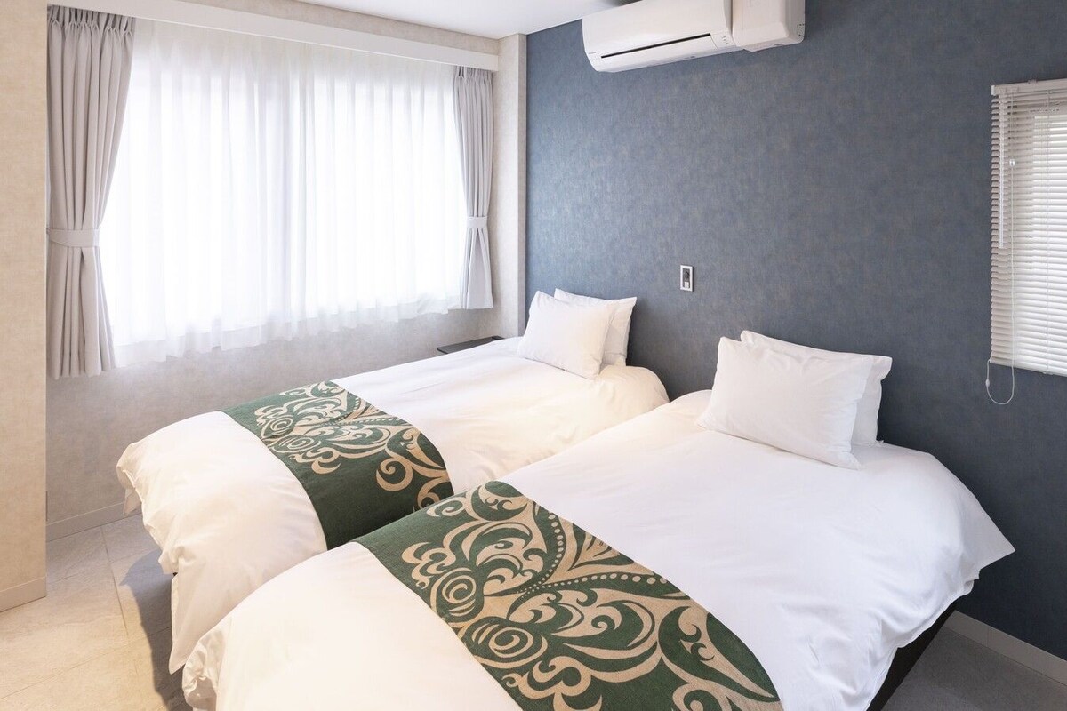 A hotel room with two beds, a window with curtains, an air conditioning unit, and a wall-mounted light switch.