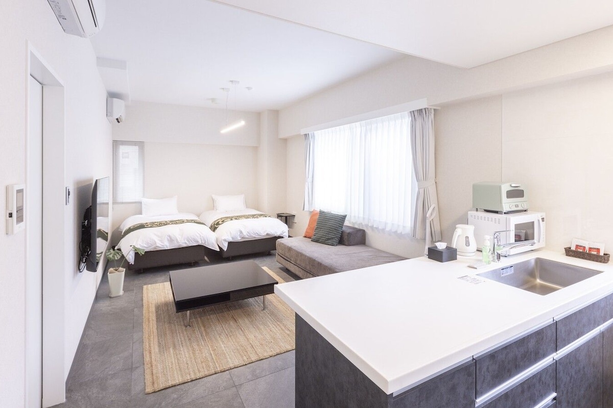 A modern hotel room with a large bed, a sofa, a dining area, and a kitchenette.
