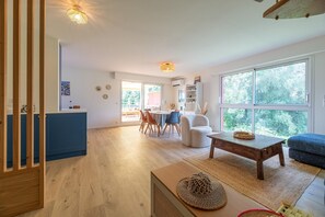 Un vaste espace de vie doté d’un plancher en bois, d’une table à manger avec des chaises, d’un canapé et d’une grande fenêtre donnant sur un paysage verdoyant.